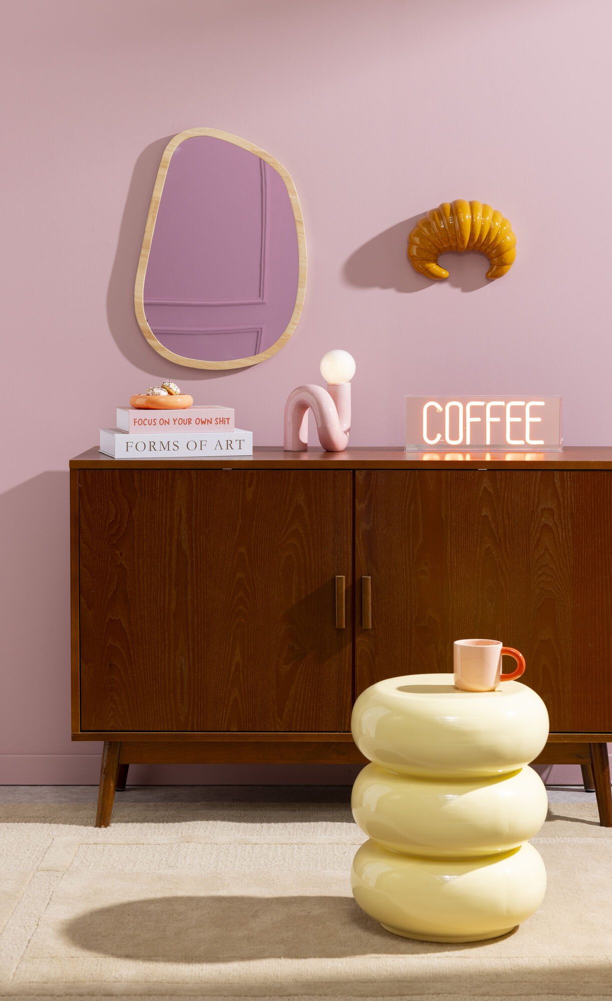 Sfeervol hoekje met roze muur, houten kast, gele bijzettafel met koffiekopje en opvallende woonaccessoires zoals een croissant aan de muur en een neon 'COFFEE'-bord